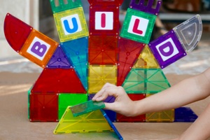 A child's hand holds a plastic magnetic tile, and in the background colourful tiles spell out build.