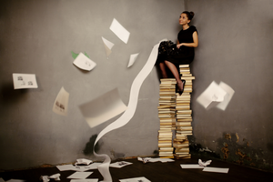 A girl sits on top of a tower of books and pages flutter around her